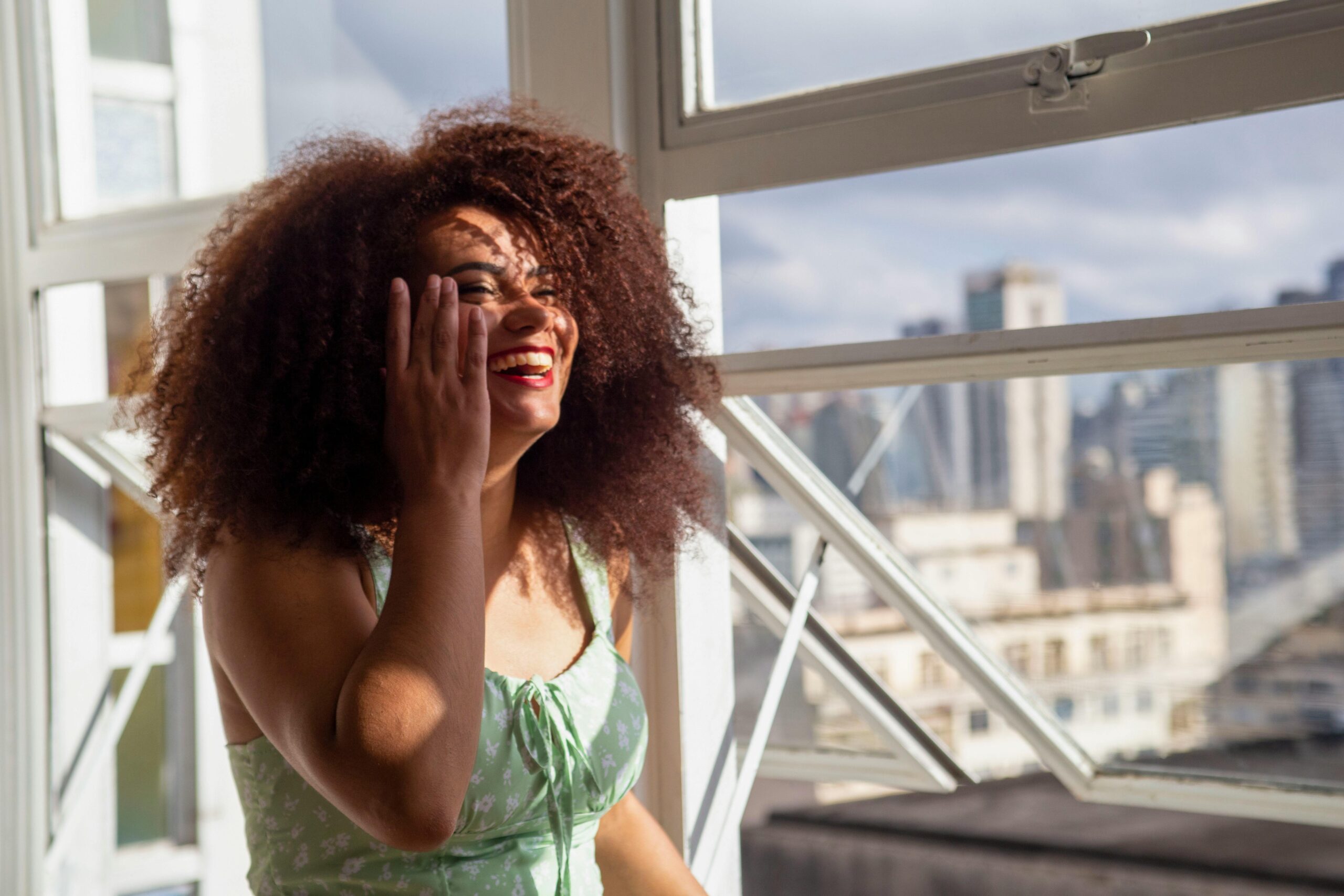 Eine fröhliche Frau mit Afrohaar genießt das Sonnenlicht im Innenbereich vor einer städtischen Kulisse.
