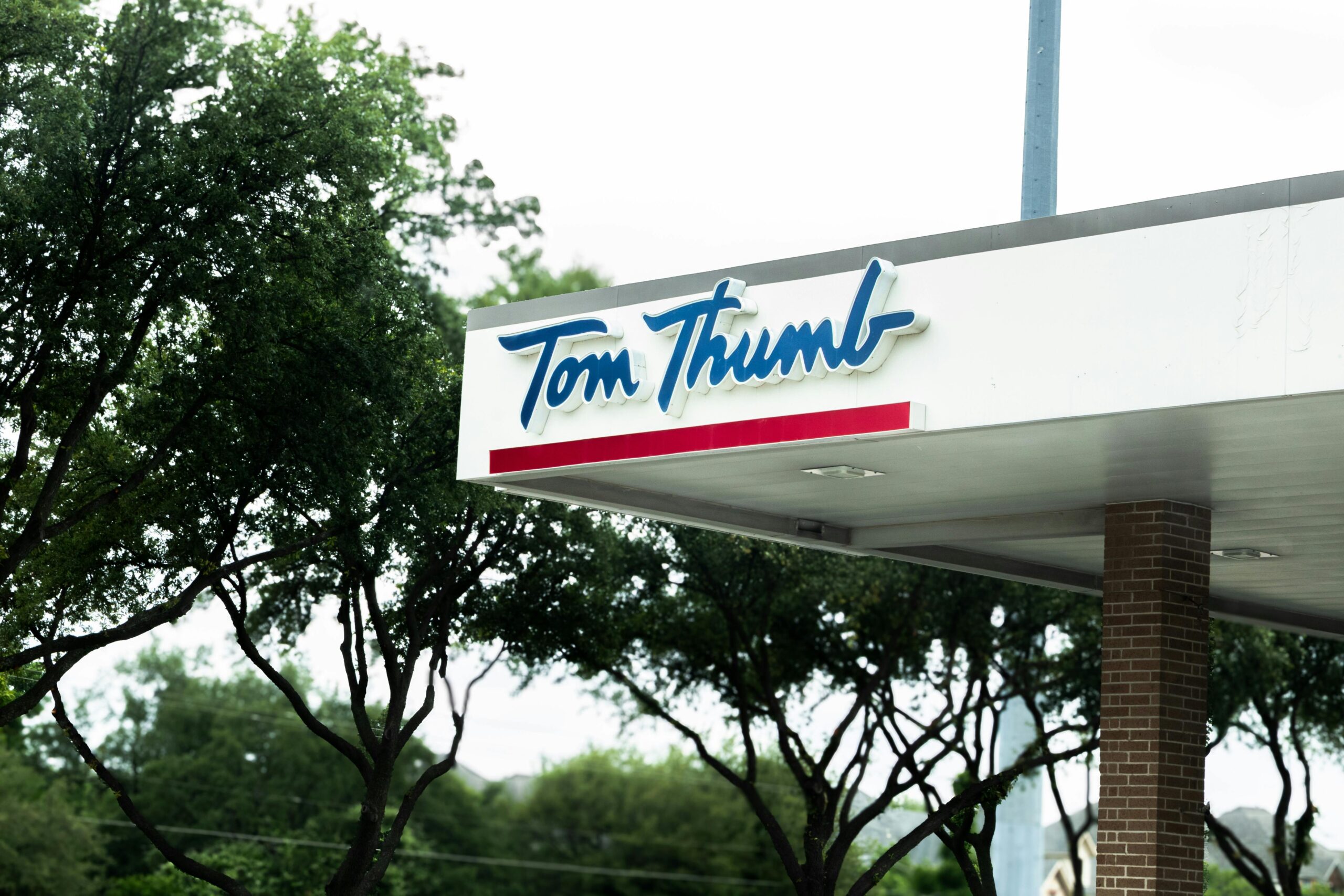 Foto des Ladenschilds von Tom Thumb mit Bäumen im Hintergrund, die eine Einzelhandelsatmosphäre im Freien vermitteln.