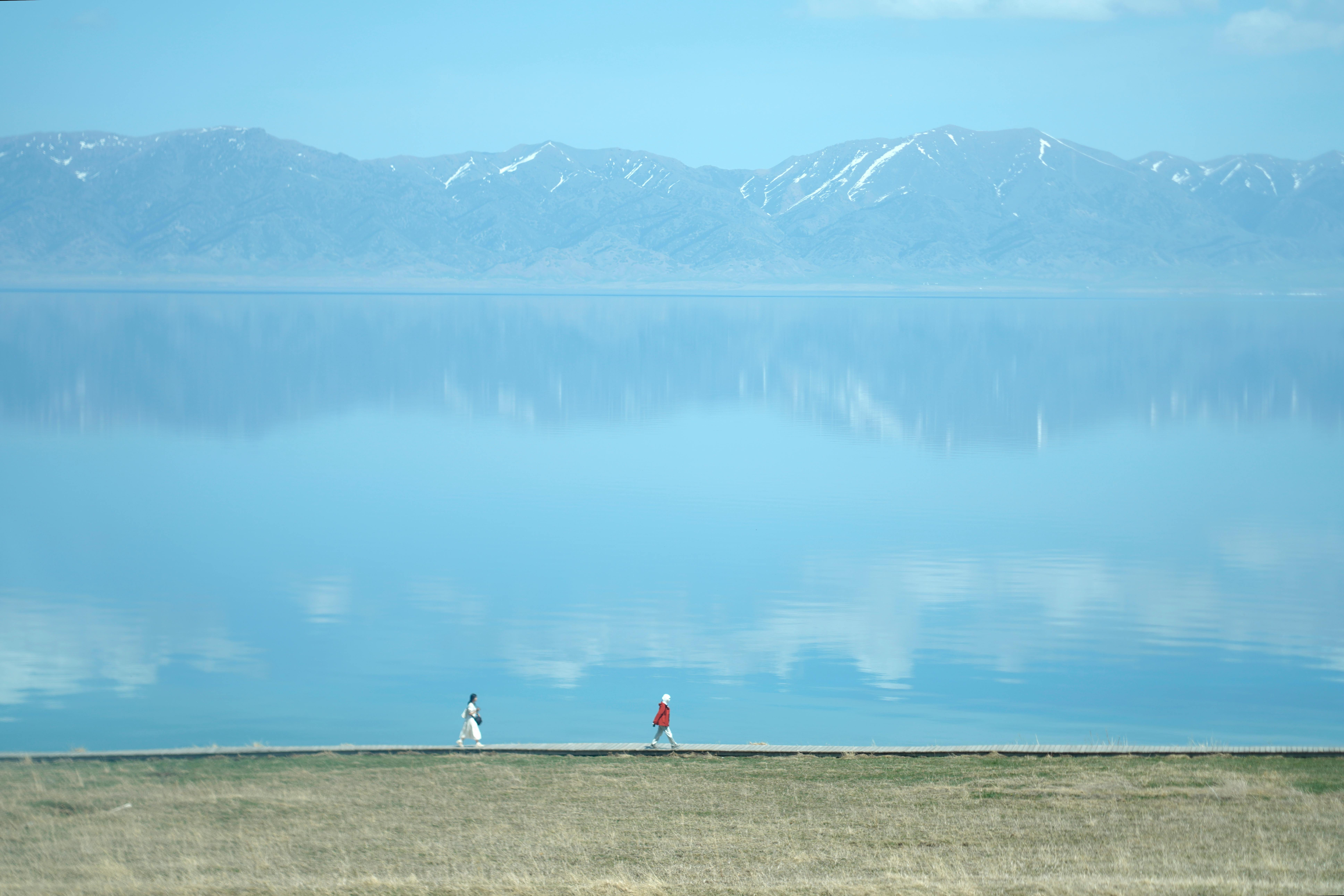 Zwei Gestalten gehen an einem ruhigen See entlang, der von schneebedeckten Bergen und blauem Himmel umgeben ist.