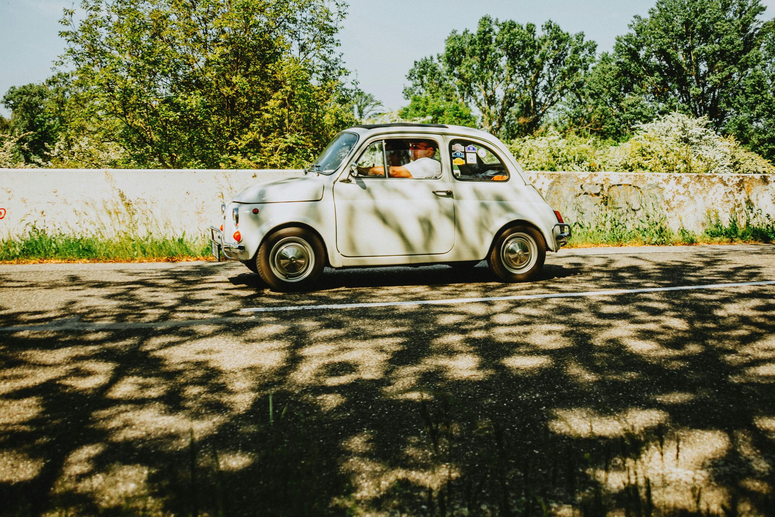 Ein klassischer Fiat 500 fährt auf einer sonnigen Straße, umgeben von üppigem Grün.