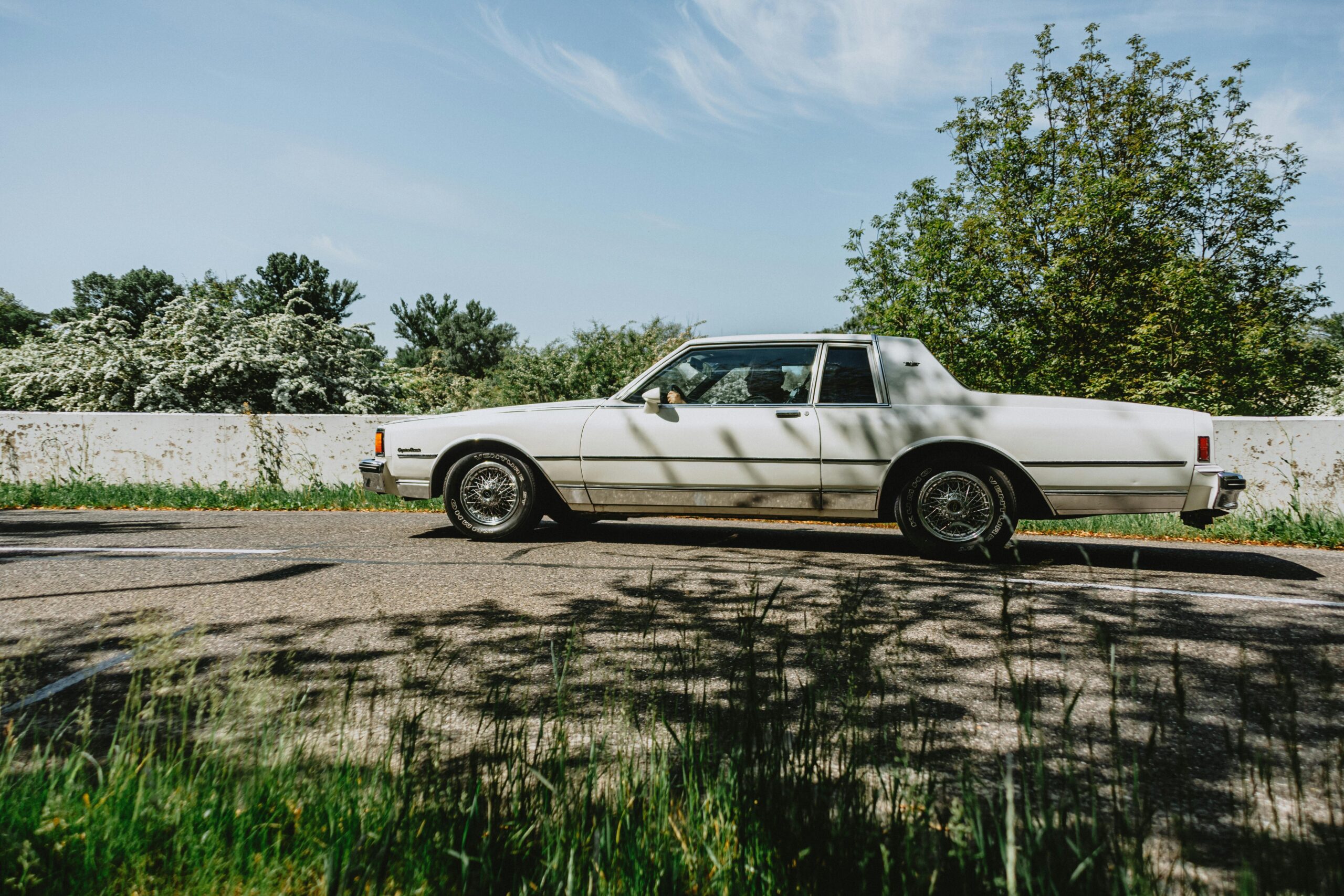 Ein weißer Oldtimer fährt auf einer sonnigen Straße, umgeben von üppigem Grün.