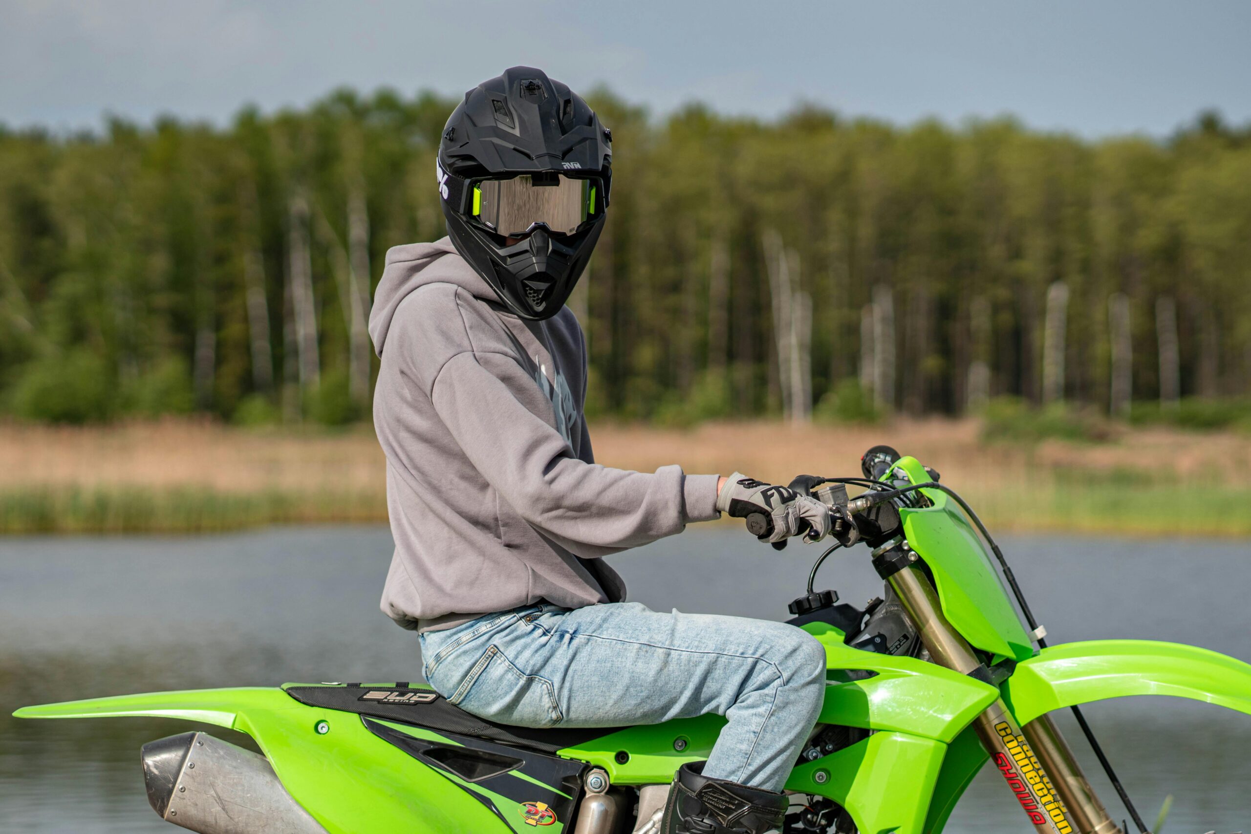 Fahrer mit Helm auf grünem Motorrad an einem See mit Wald im Hintergrund.
