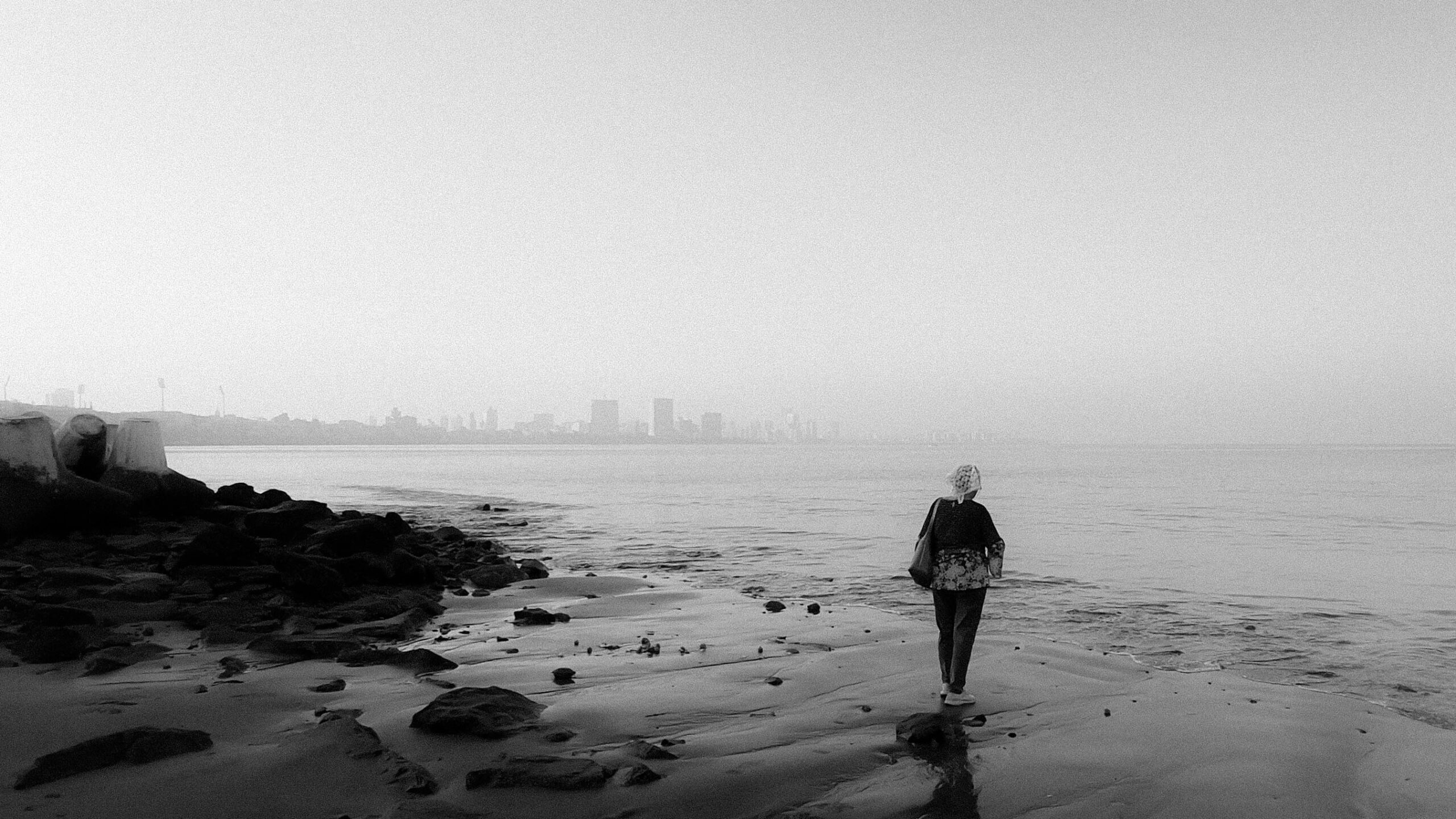 Schwarzweißfoto einer Frau, die an einem felsigen Strand entlanggeht und in der Ferne die Skyline einer Stadt vor Augen hat.