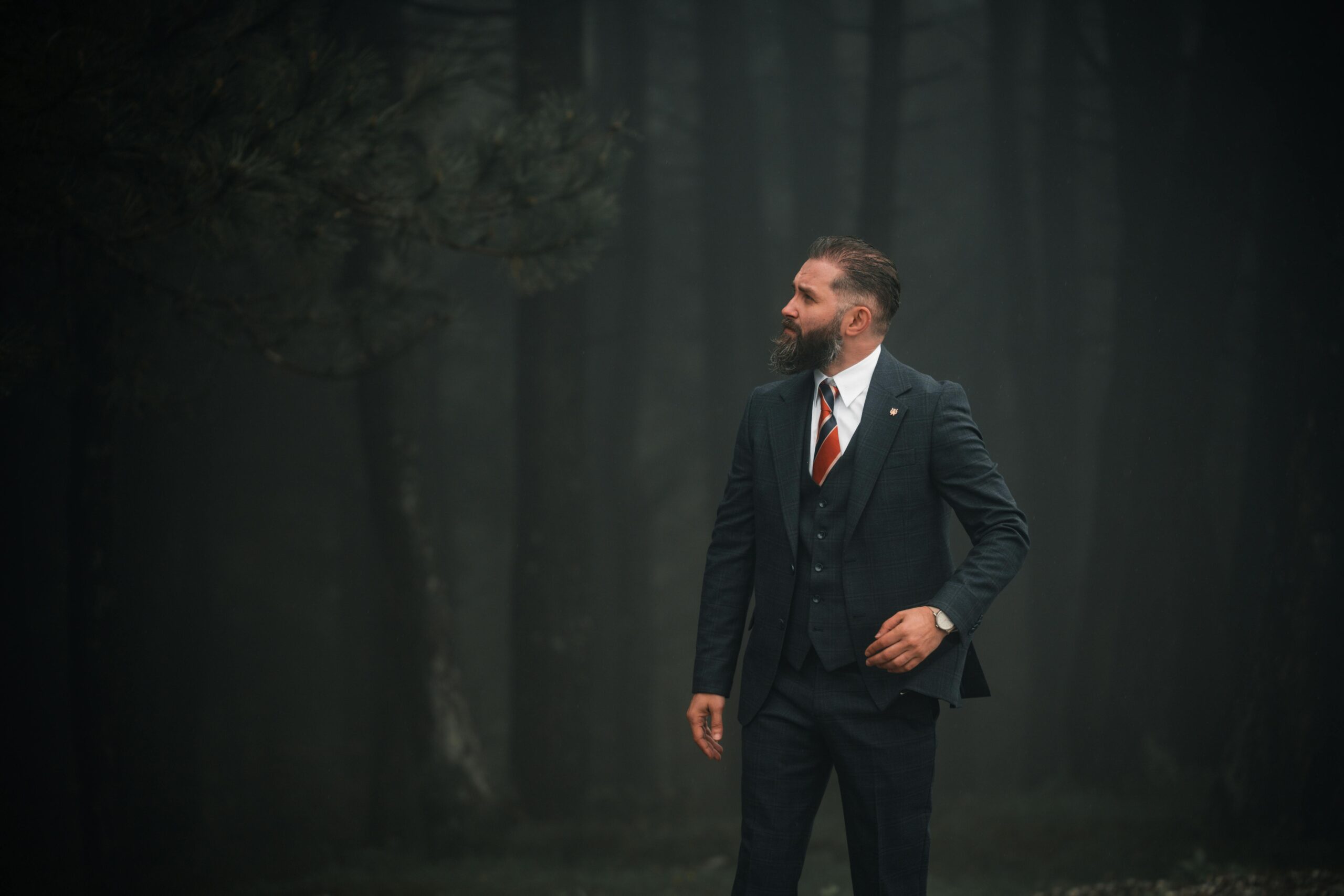 Ein Mann im Anzug steht selbstbewusst in einem nebligen Wald und strahlt Eleganz aus.