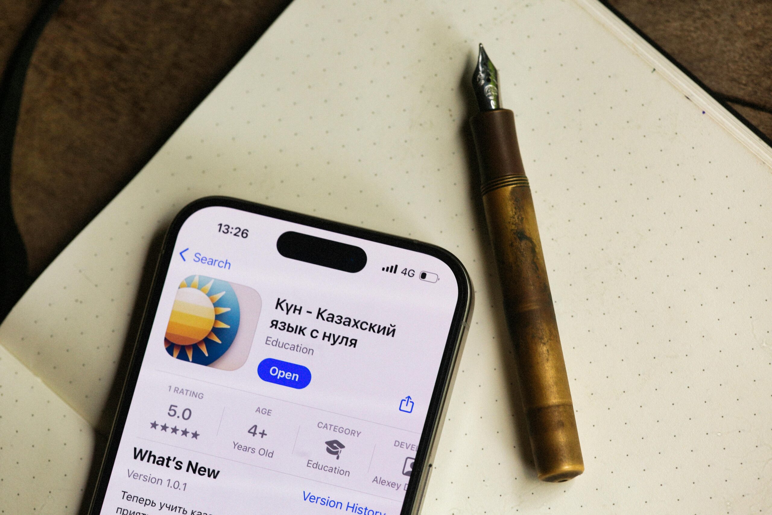 Smartphone mit kasachischer Sprach-App neben einem Füllfederhalter auf einem Notizbuch.