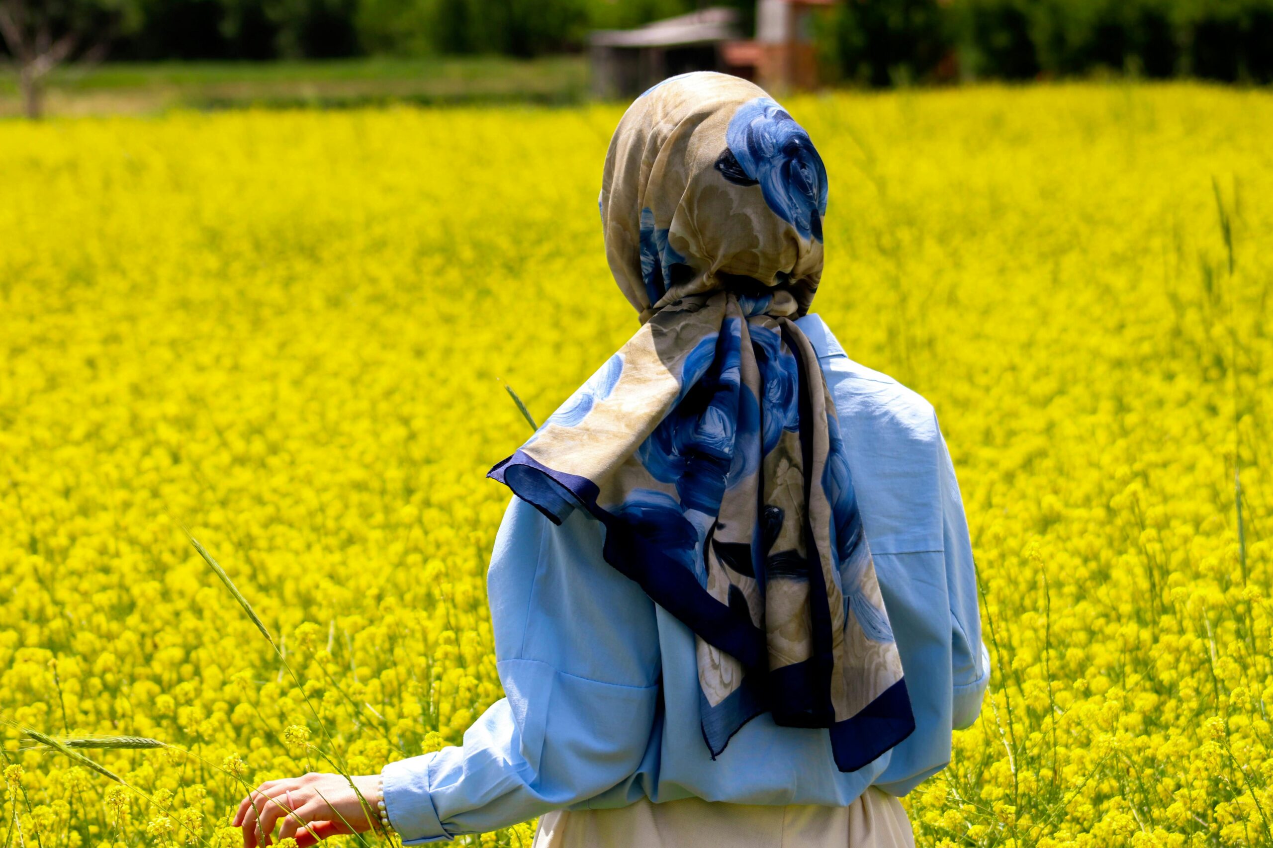 Eine Frau mit einem geblümten Schal und einem blauen Hemd geht unter einem sonnigen Himmel durch ein leuchtend gelbes Feld.