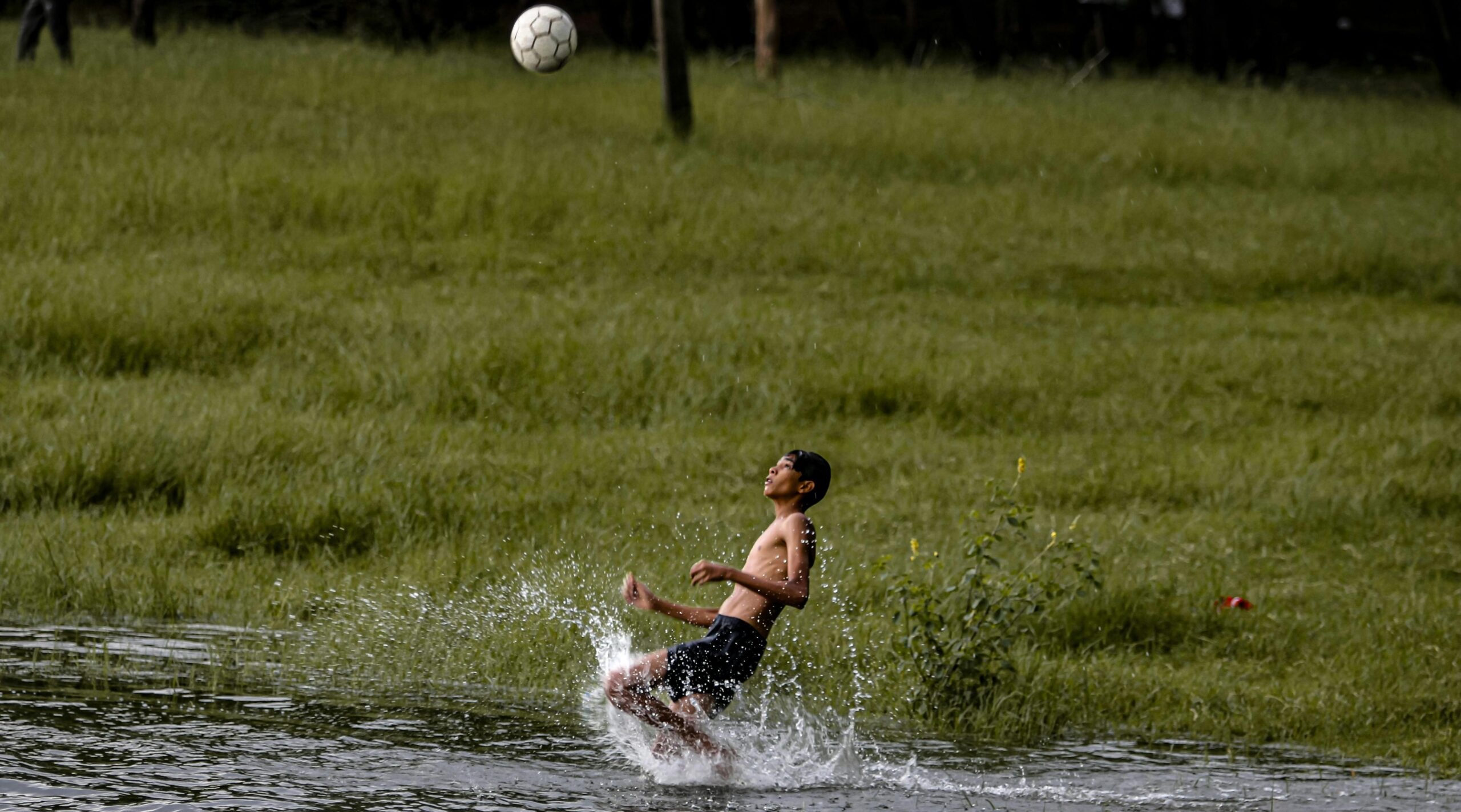 Ein kleiner Junge kickt einen Fußball, während er im Wasser planscht, vor der Kulisse eines üppigen grünen Feldes.