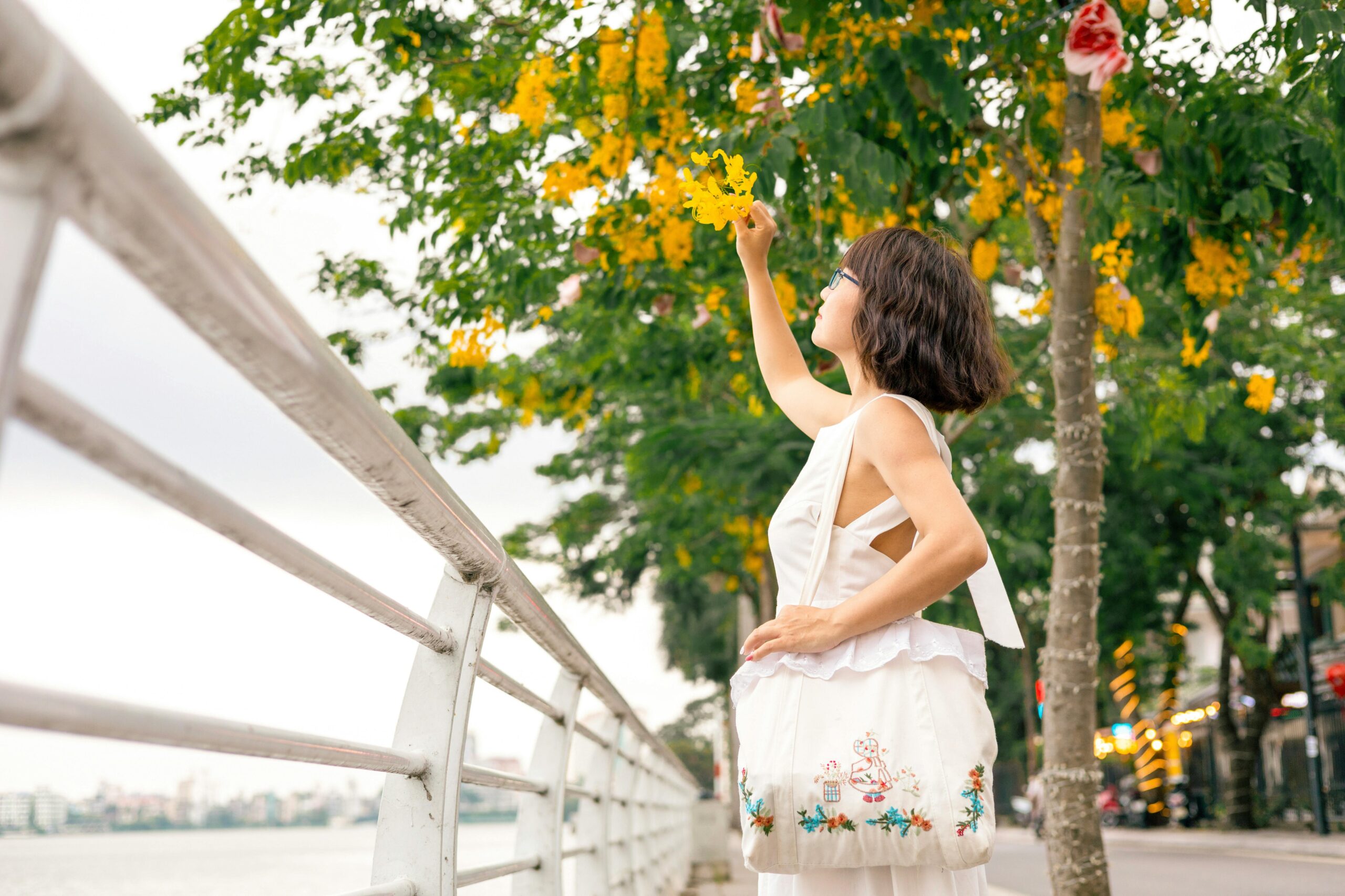 Eine Frau in einem weißen Kleid pflückt leuchtend gelbe Blumen entlang eines Weges an einem See.