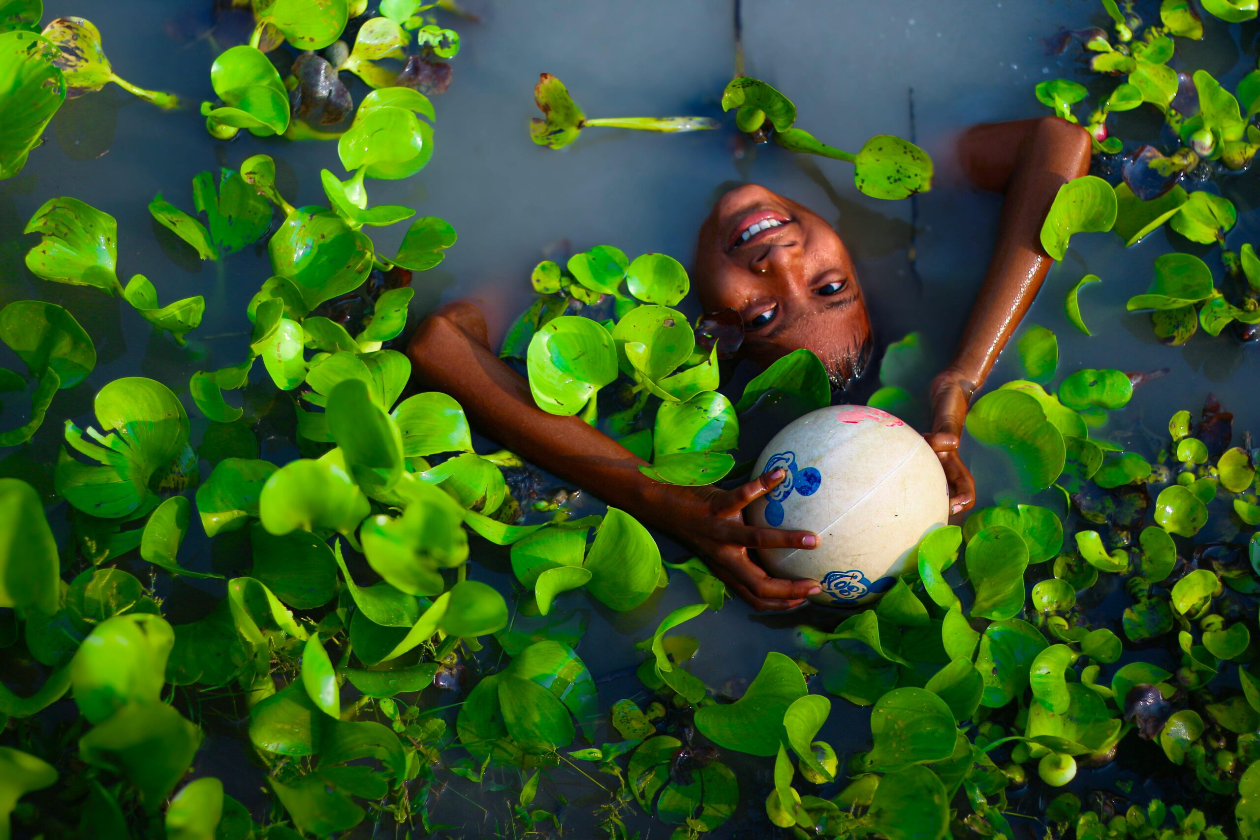 Ein Kind spielt fröhlich im Wasser, umgeben von leuchtenden Wasserpflanzen in Westbengalen, Indien.