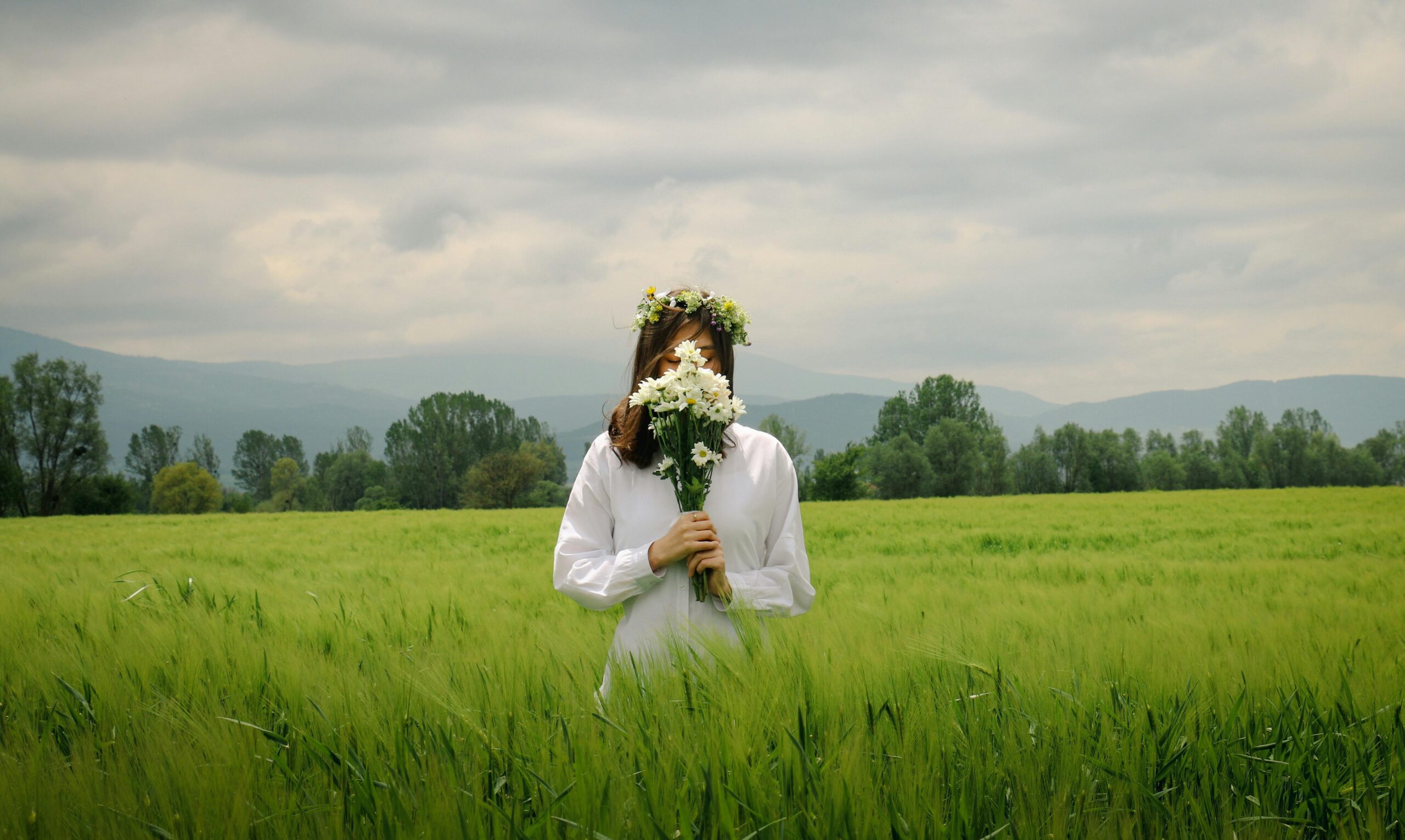 Frau in Weiß, mit Gänseblümchen in der Hand, auf einem ruhigen grünen Feld, trägt eine Blumenkrone.