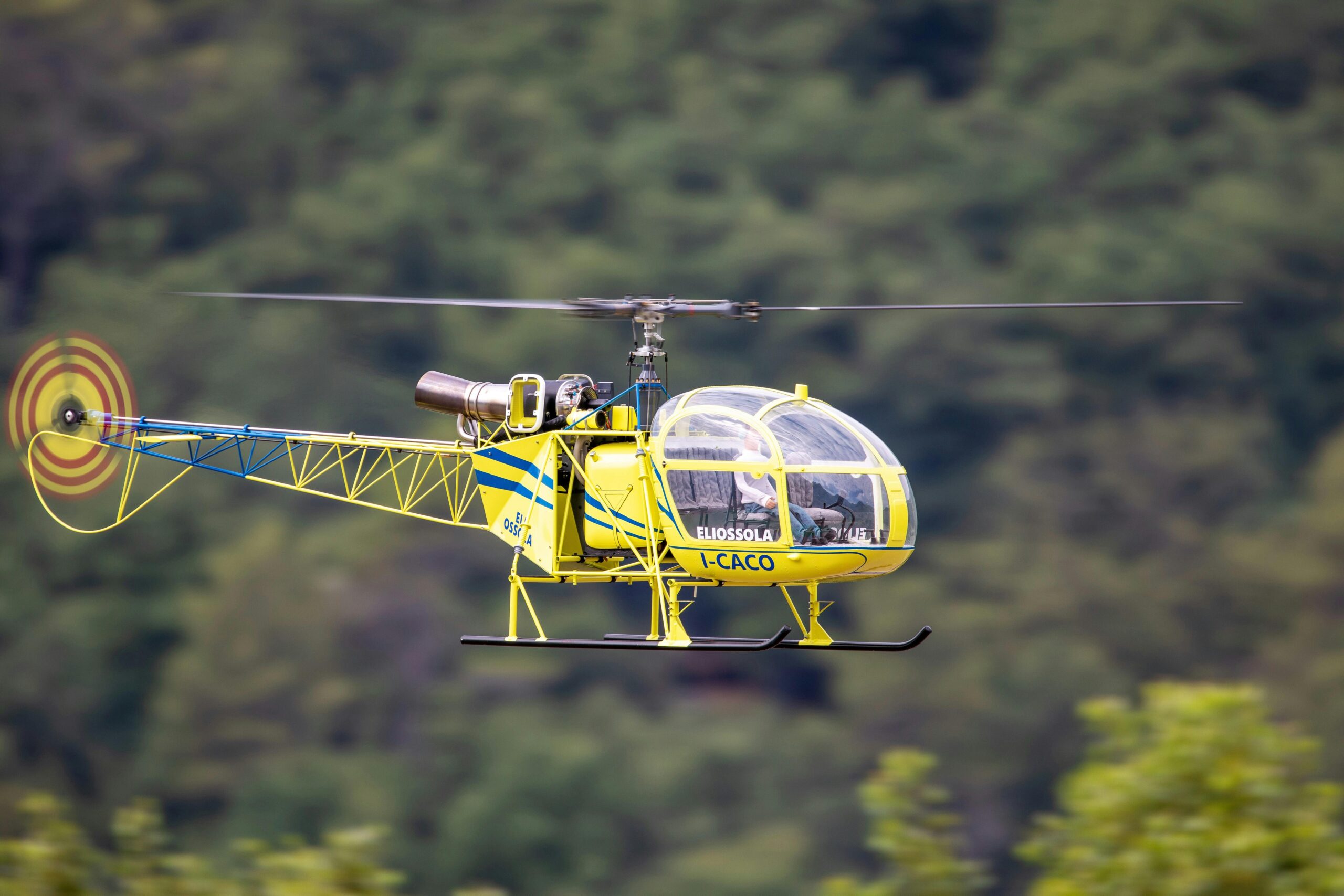 Ein leuchtend gelber Hubschrauber im Flug vor einem verschwommenen Waldhintergrund, der dynamische Bewegungen zeigt.