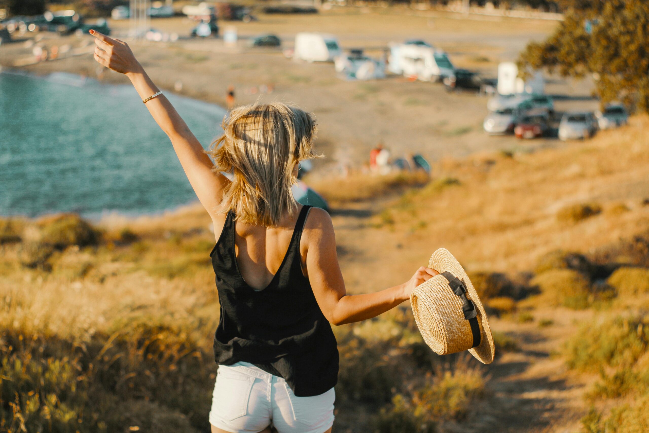 Blonde Frau im schwarzen Oberteil hebt freudig den Arm in der Nähe eines Strandes mit malerischer Kulisse.