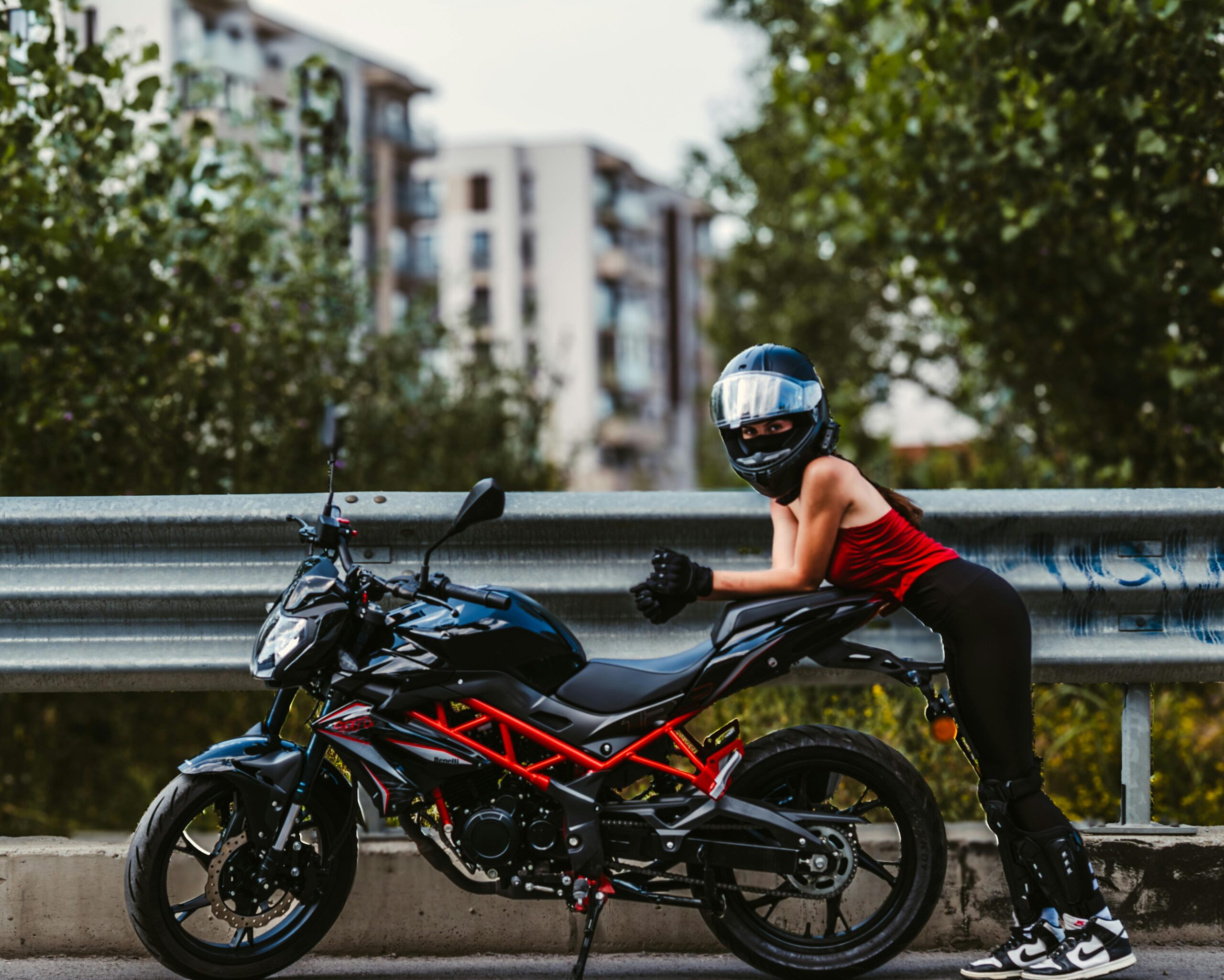 Eine Motorradfahrerin in Montur posiert neben einem Motorrad am Straßenrand einer Stadt und präsentiert die moderne Motorradkultur.