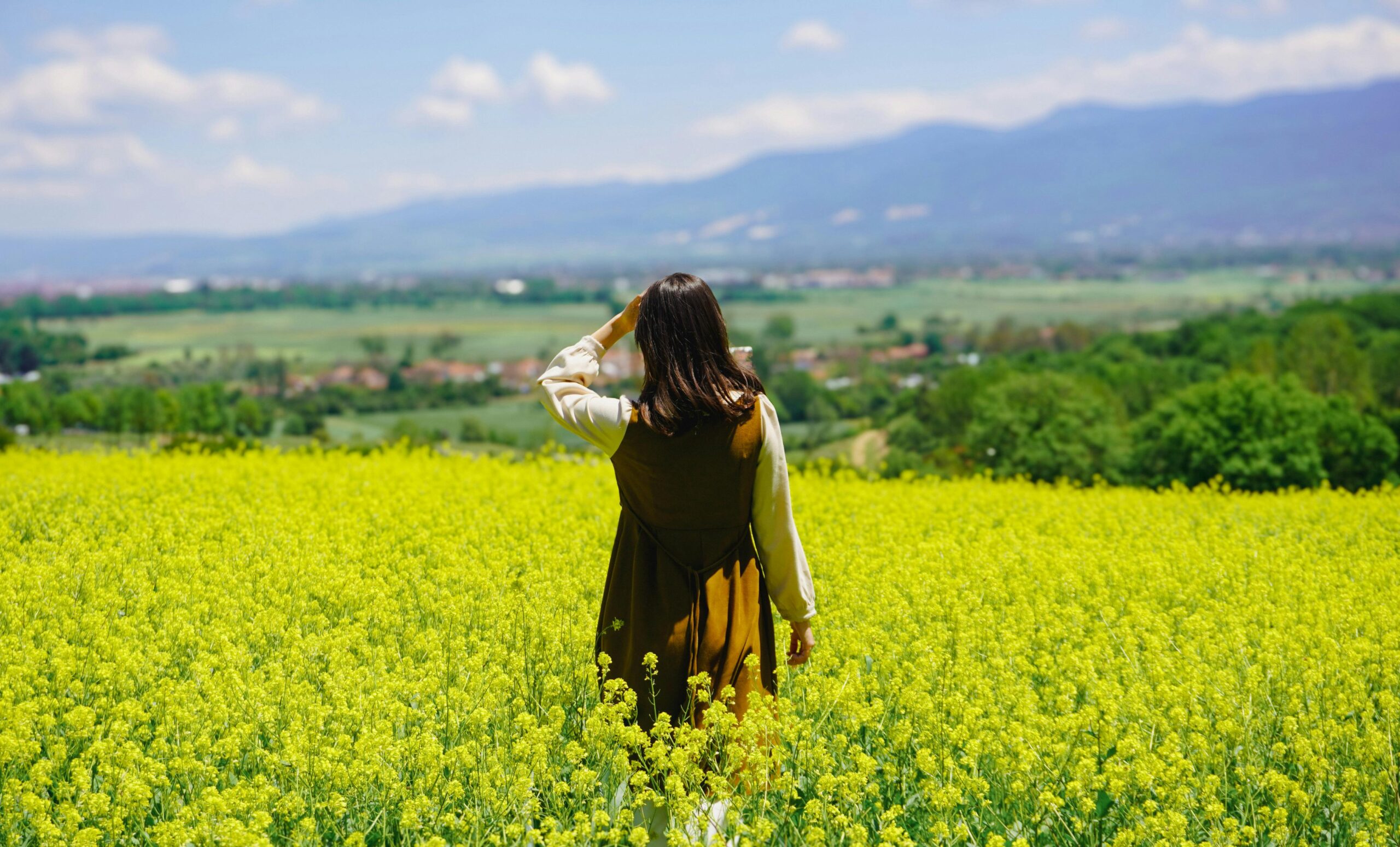 Frau steht an einem Sommertag auf einem leuchtend gelben Blumenfeld und blickt auf die malerischen Berge.
