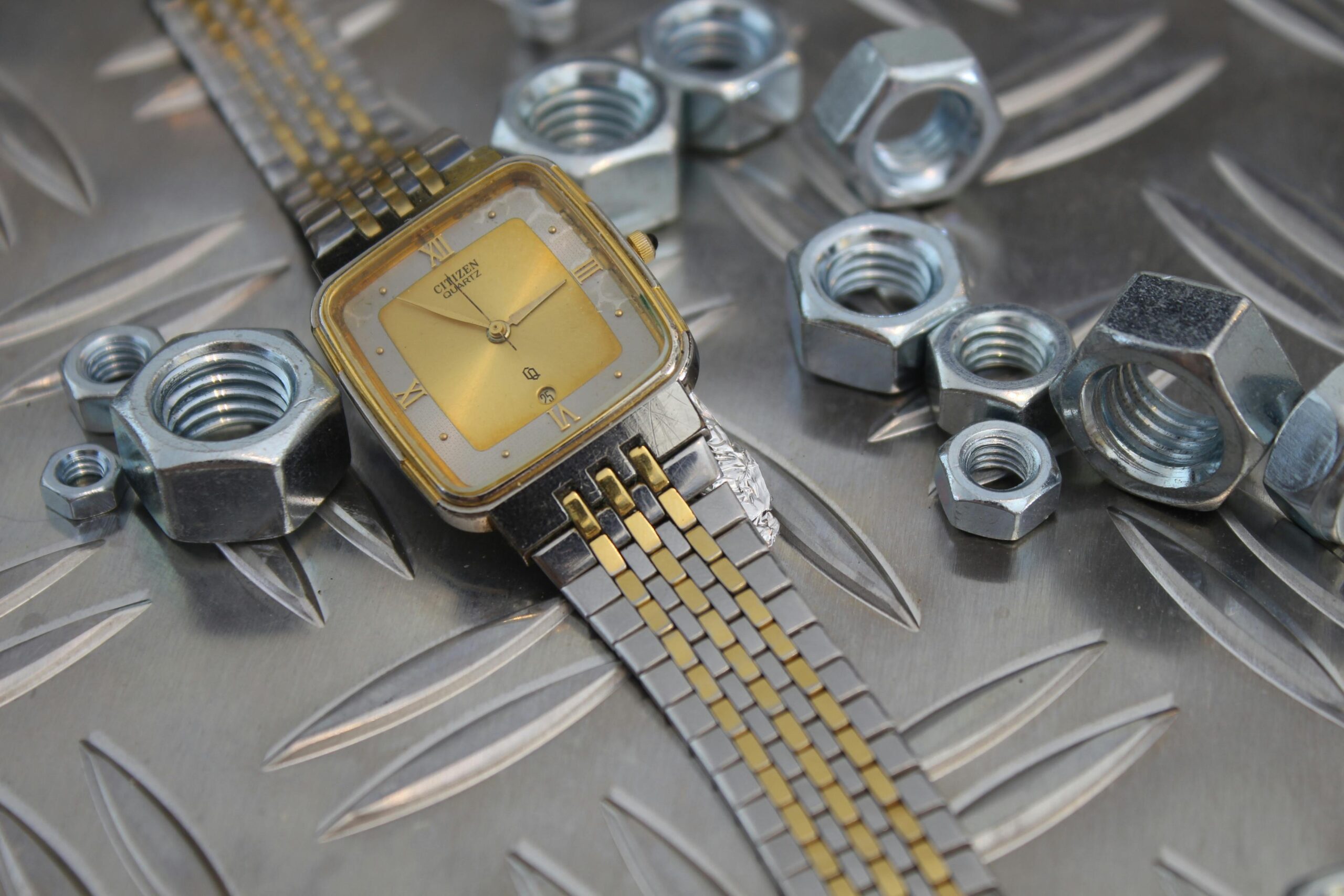 Eine stilvolle Uhr in Gold und Silber auf einer industriellen Stahloberfläche mit Metallmuttern drumherum.