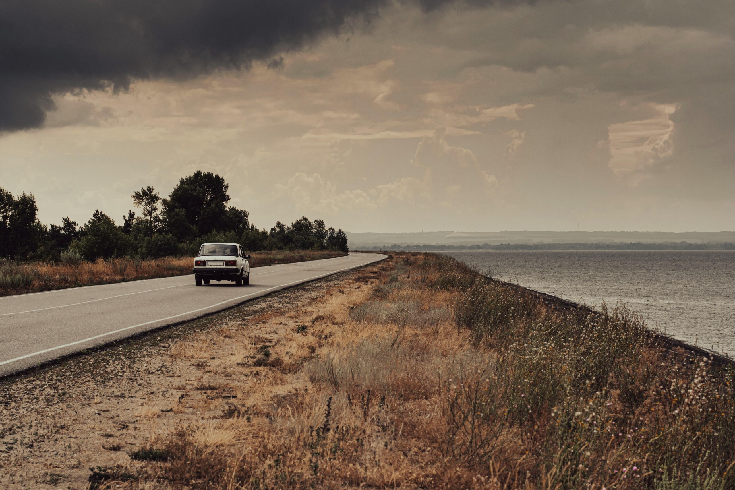 Ein Oldtimer fährt in der Dämmerung unter dramatischem Himmel eine verlassene Küstenstraße entlang.