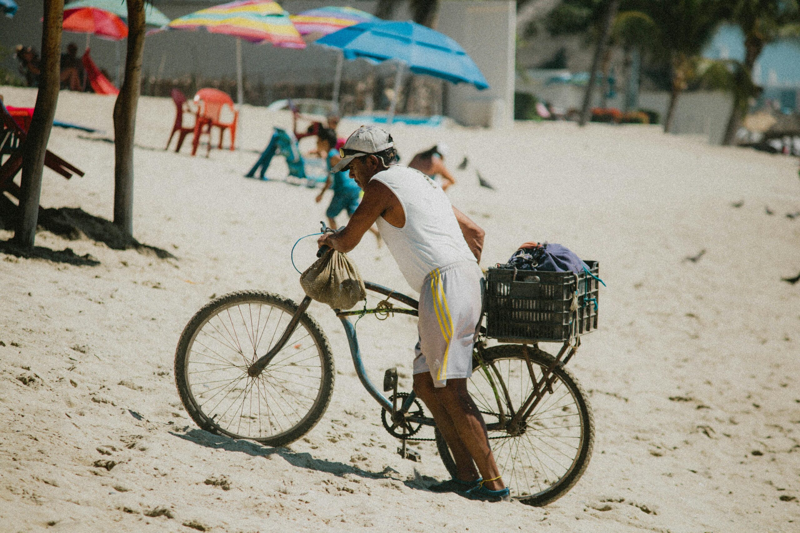 Ein Mann schiebt ein Fahrrad an einem sonnigen Strand mit bunten Sonnenschirmen im Hintergrund.