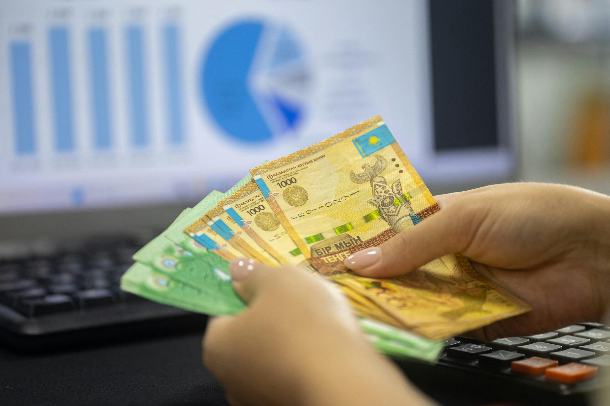 Hände halten kasachische Tenge-Scheine mit Finanzdiagrammen und Grafiken auf dem Computerbildschirm.