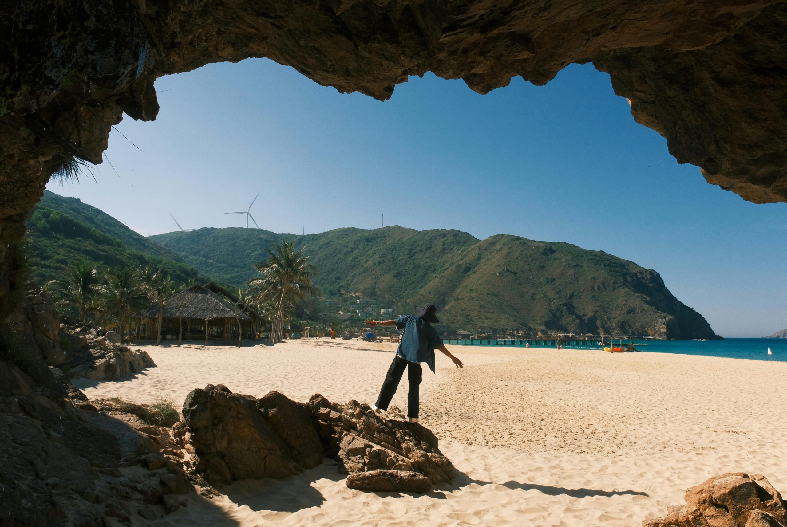 Person erkundet einen Sandstrand mit Bergen und Windturbinen im Hintergrund.