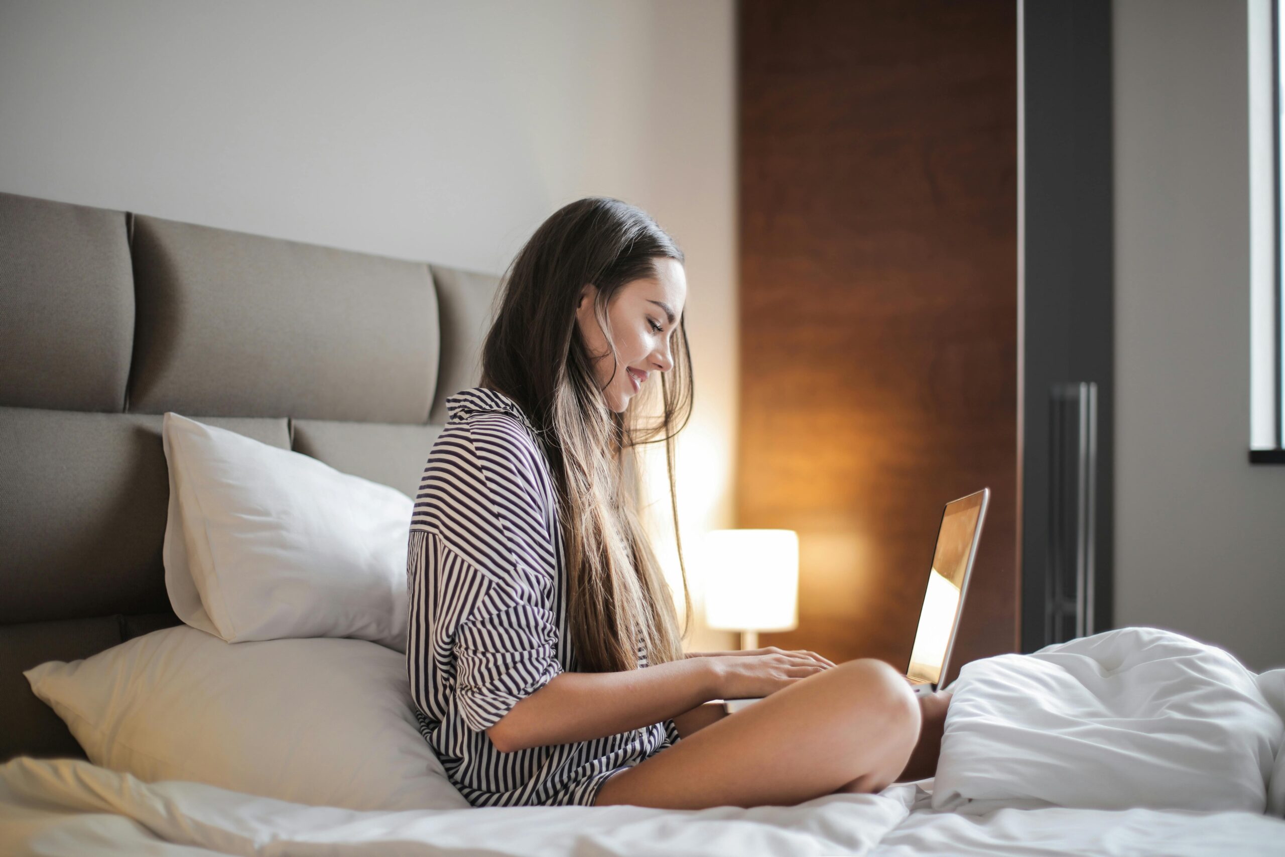 Seitenansicht Foto Der Lächelnden Frau In Einem Schwarz Weiß Gestreiften Oberteil, Das Auf Einem Bett Sitzt, Während Man Einen Laptop Benutzt