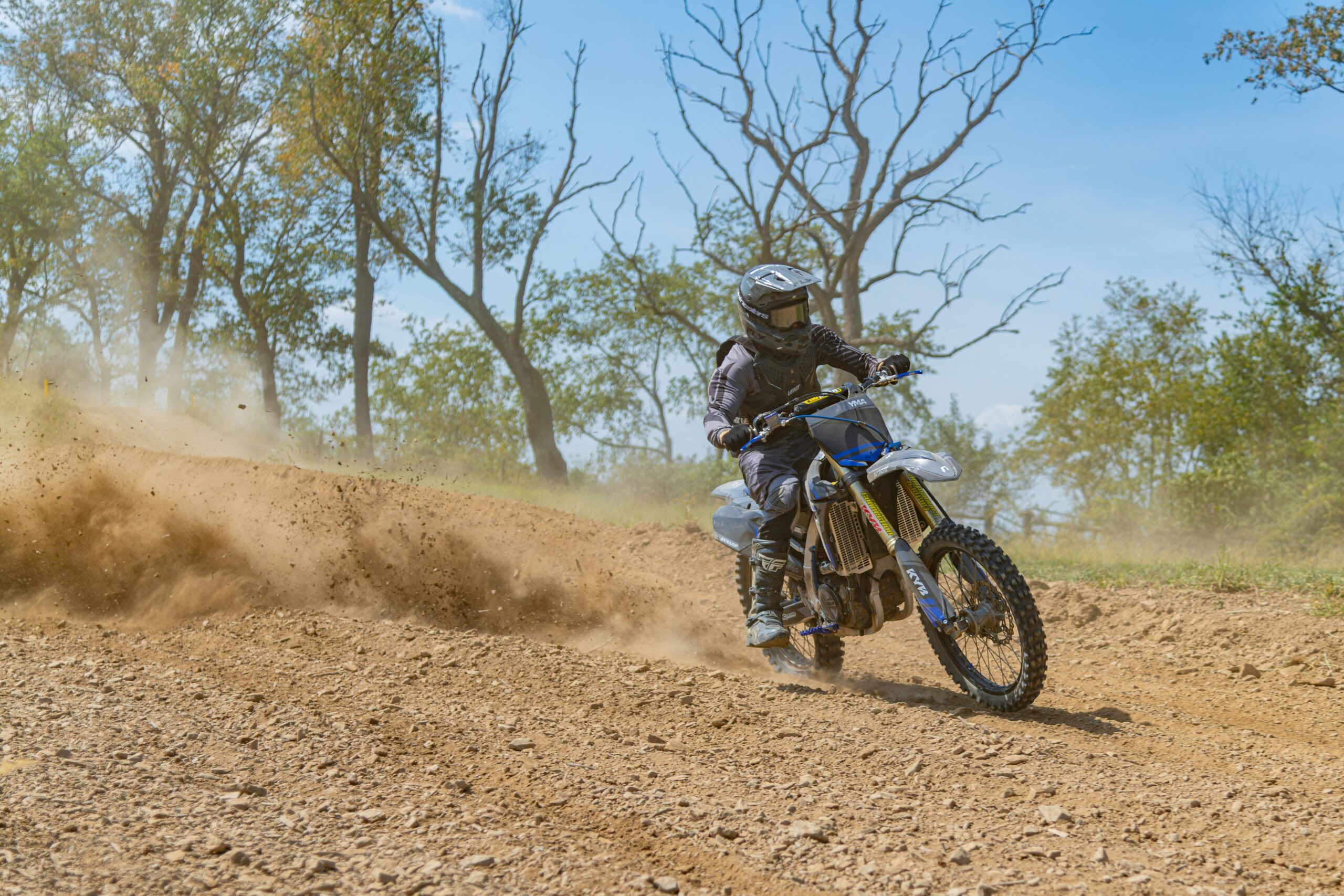 Aufregende Dirtbike-Fahrt auf einer holprigen Feldstrecke im Freien. Staub fliegt, Bäume im Hintergrund.