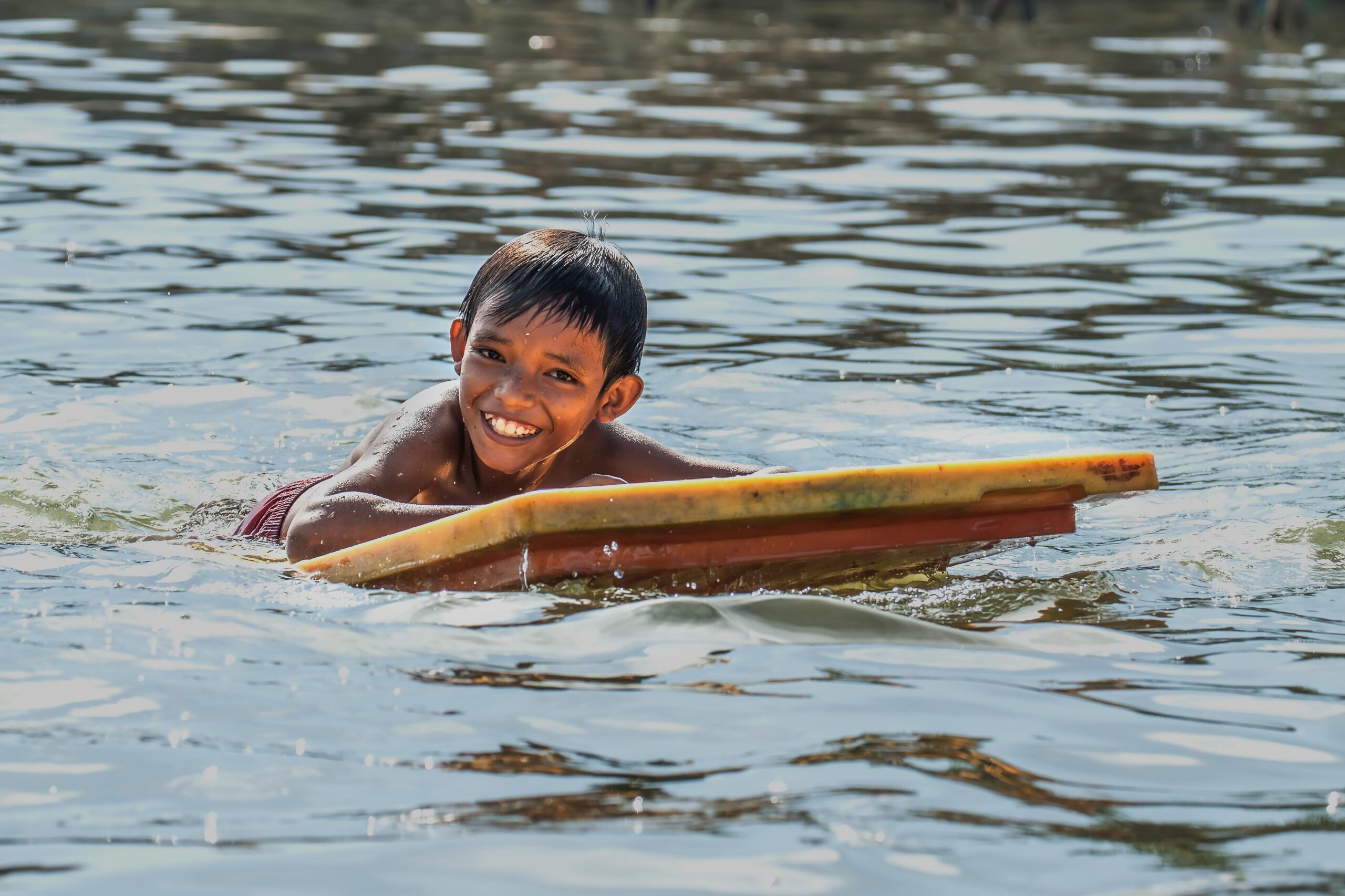 Ein glücklicher Junge schwimmt in einem See, während er sich an einem Holzbrett festhält, und genießt einen sonnigen Tag.