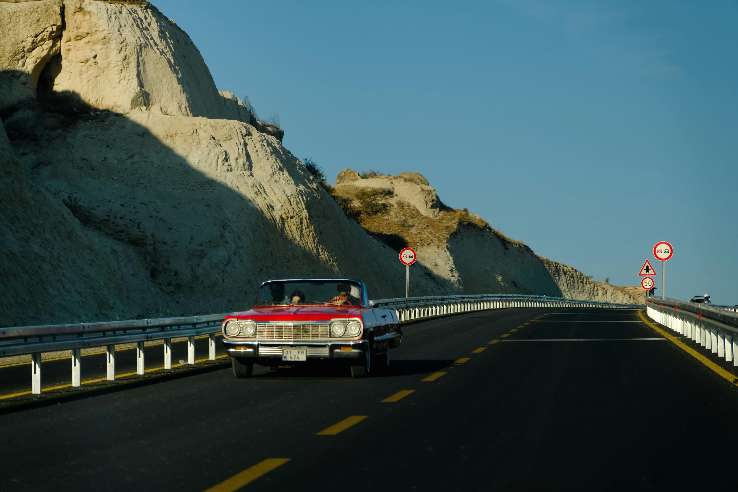 Ein rotes Oldtimer-Cabrio fährt auf einer malerischen Autobahn mit felsigen Klippen unter einem klaren blauen Himmel.