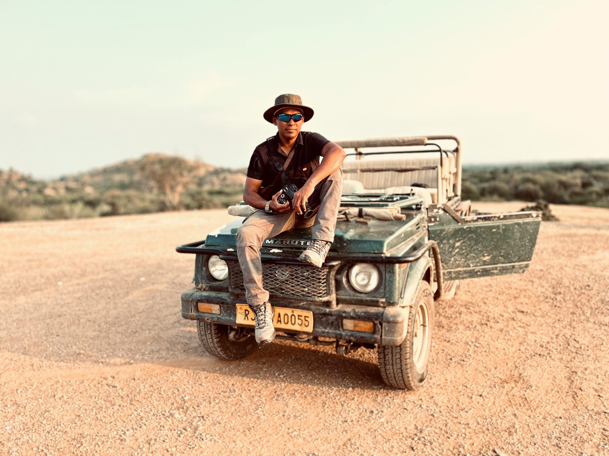 Ein Mann mit Sonnenbrille sitzt auf einem Jeep in einer trockenen, rauen Landschaft und strahlt Abenteuerlust aus.