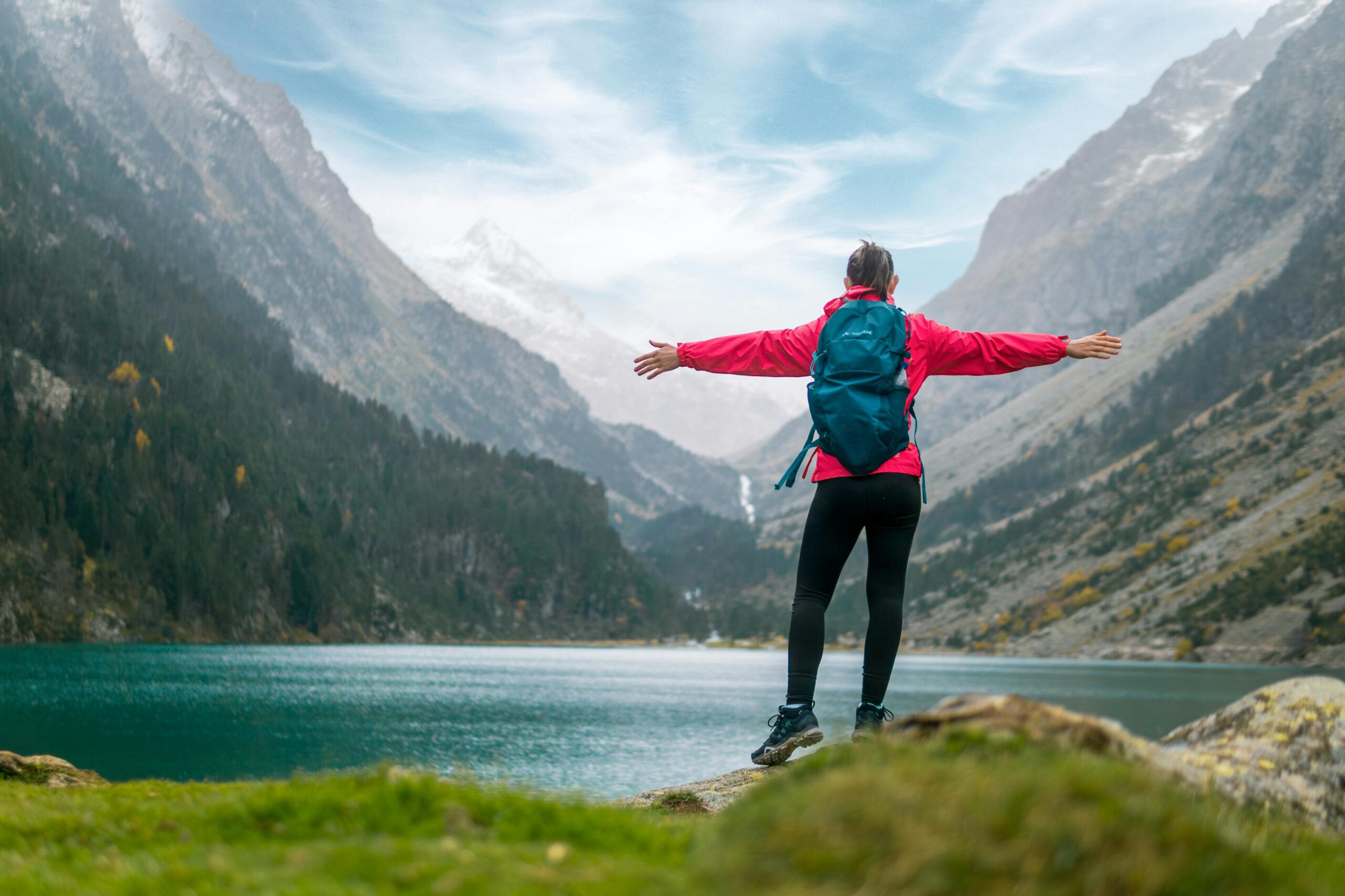 Eine Wanderin in einer roten Jacke steht mit weit ausgebreiteten Armen da und blickt auf einen ruhigen Alpensee.