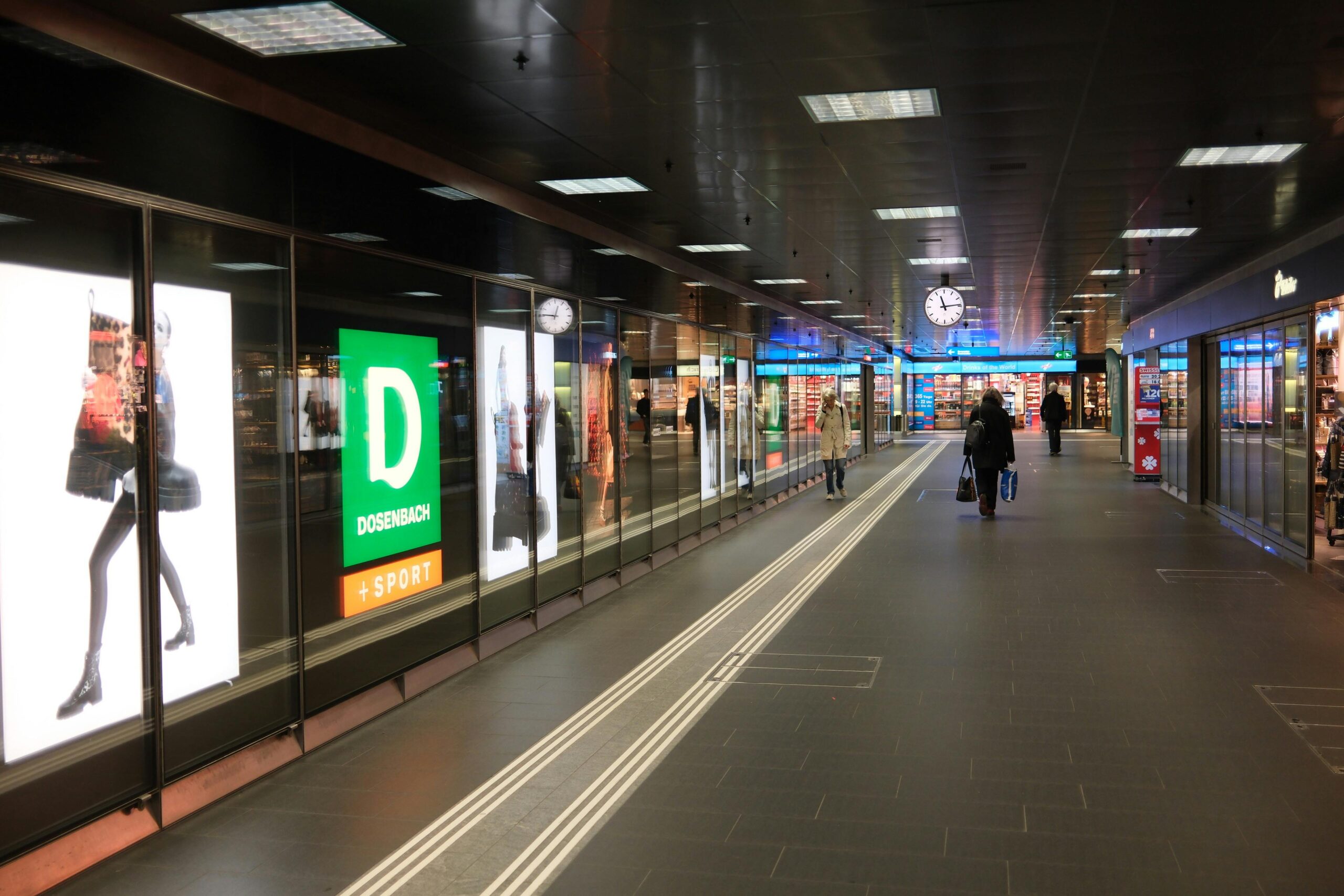 Weite Ansicht eines belebten Korridors eines Einkaufszentrums mit Menschen, Geschäften und einer markanten Uhr.