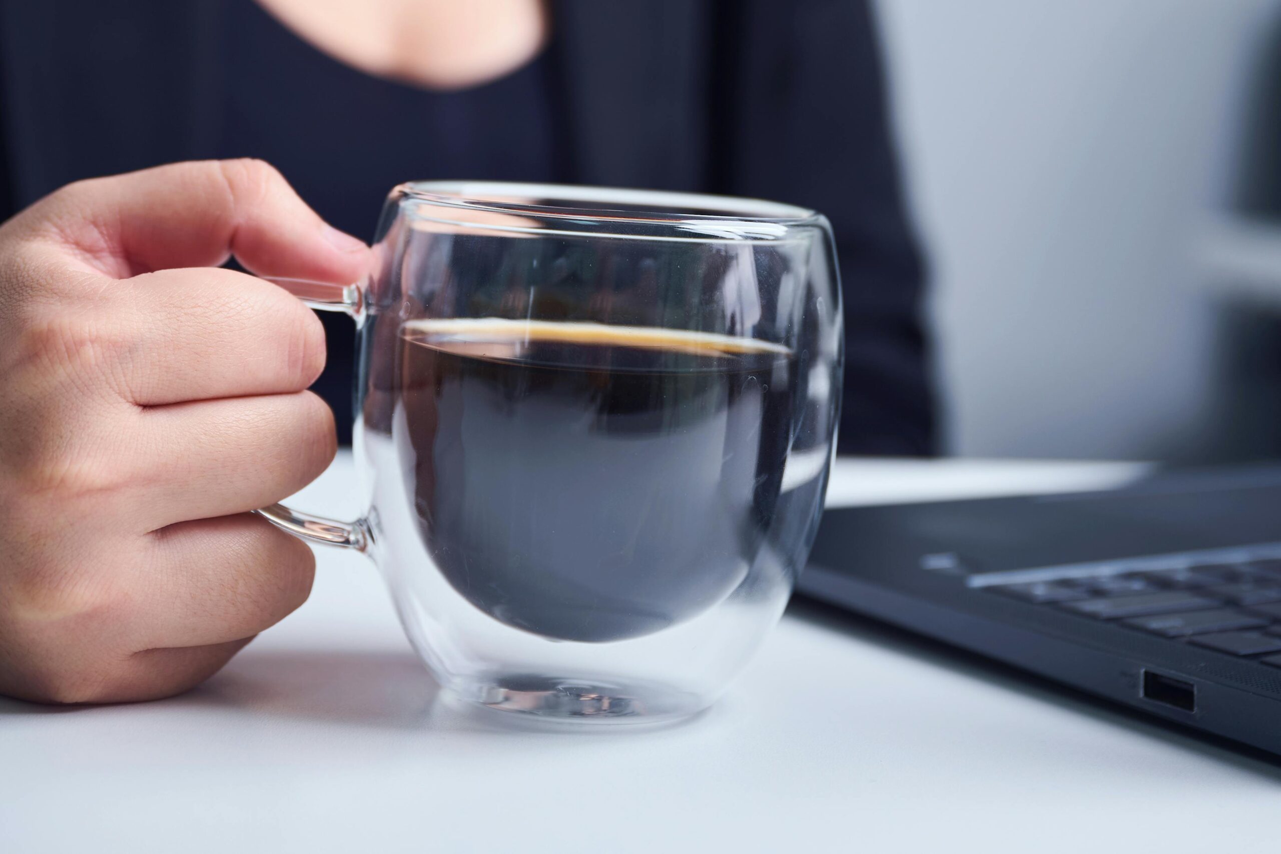 Nahaufnahme einer Hand, die eine Kaffeetasse neben einem Laptop auf einem Schreibtisch im Innenbereich hält.