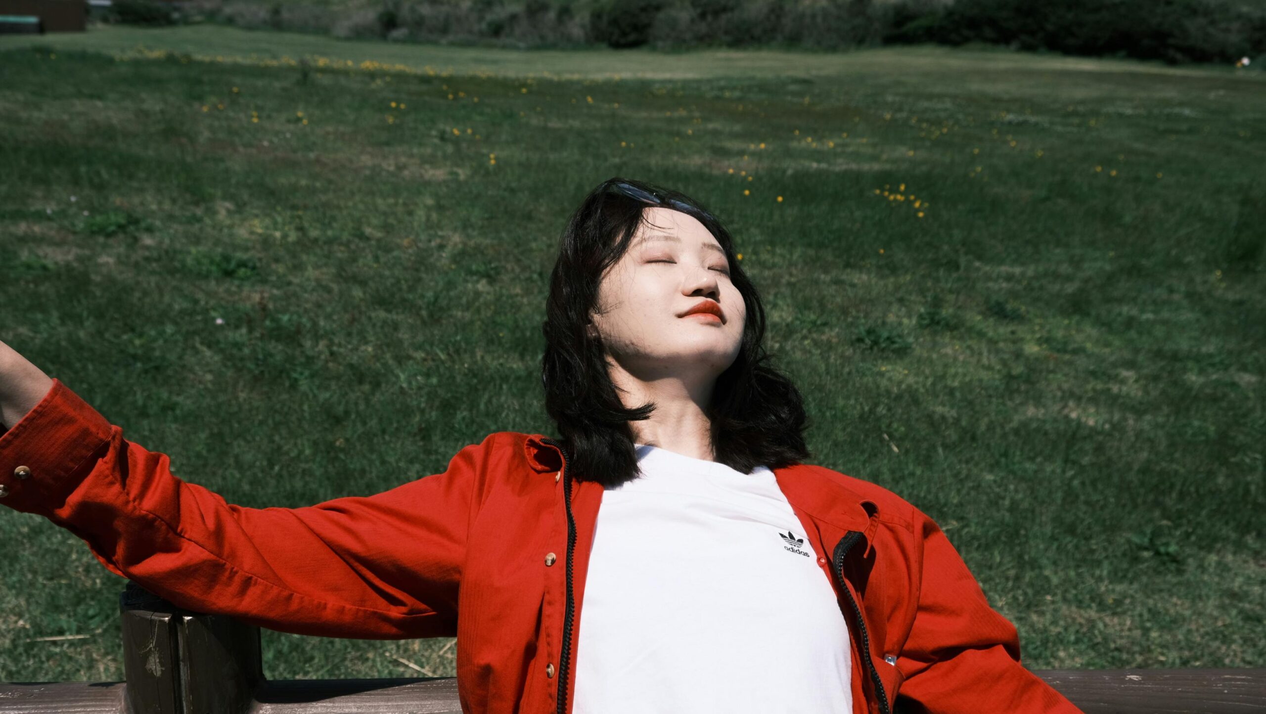 Frau genießt das Sonnenlicht im Freien in einem Park und trägt eine rote Jacke.