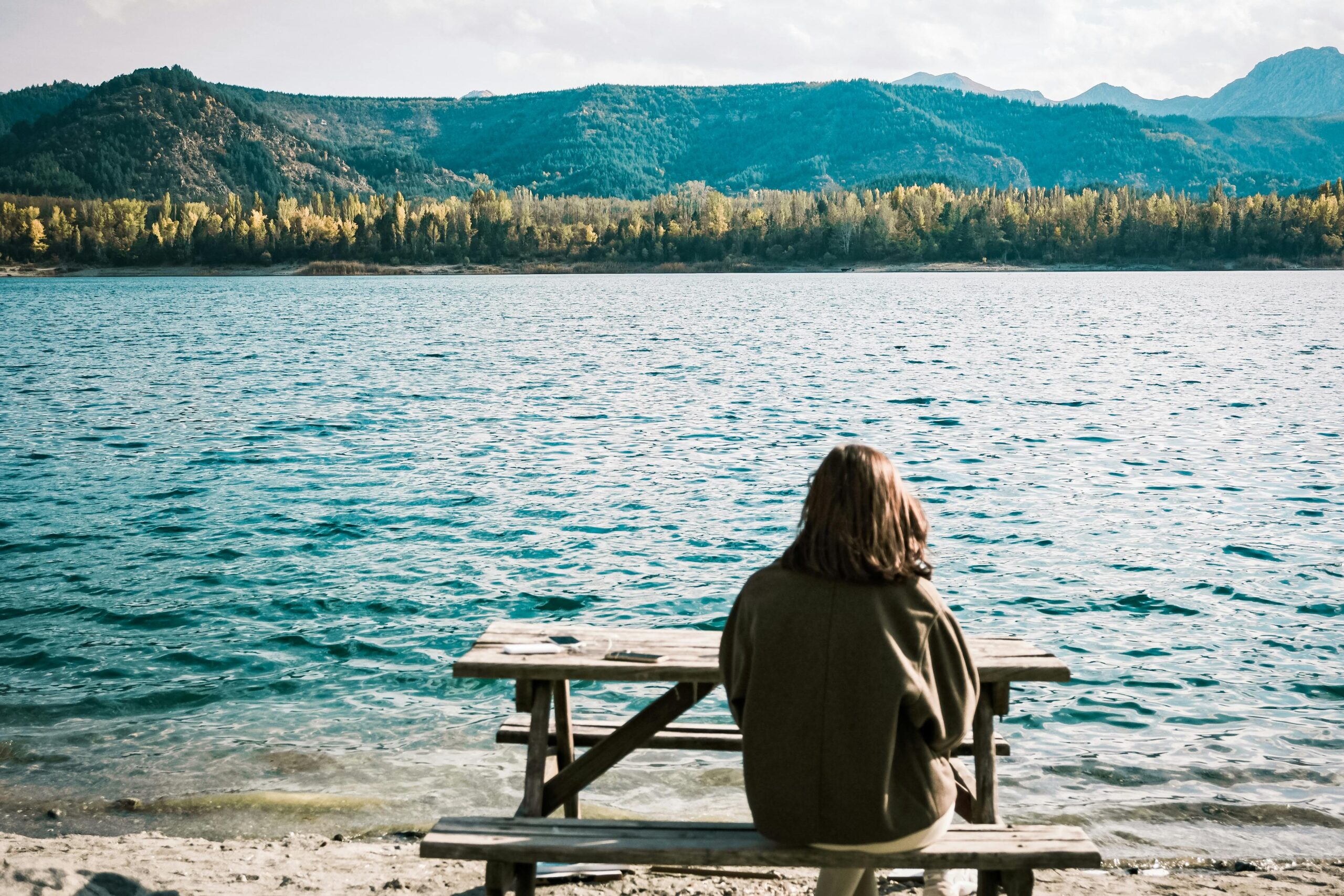 Frau sitzt auf einer Picknickbank an einem See mit einem Bergwald im Hintergrund.