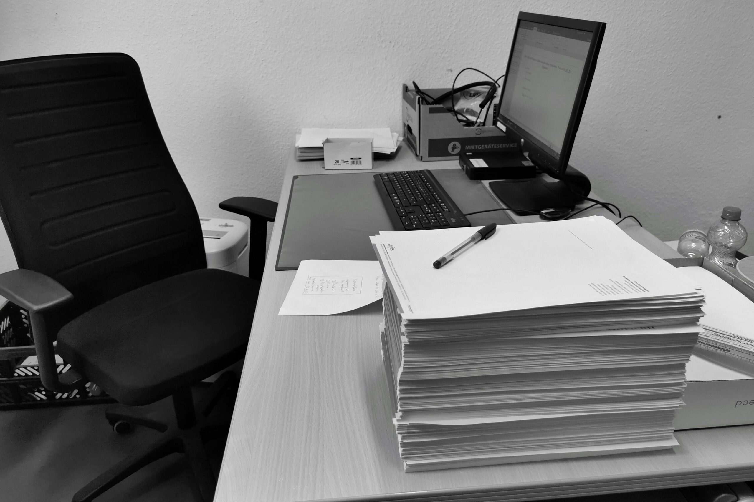 Schwarzweißbild eines überfüllten Büroschreibtischs mit Computer, Stuhl und Papierkram.